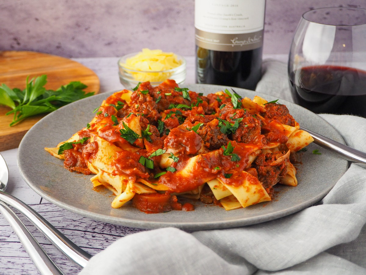 Slow cooker lamb ragu 4 Plate of slow cooker lamb ragu, garnished with chopped parsley, surrounded by serving cutlery, glass of red wine, bowl of parmesan cheese and chopping board with parsley on it.