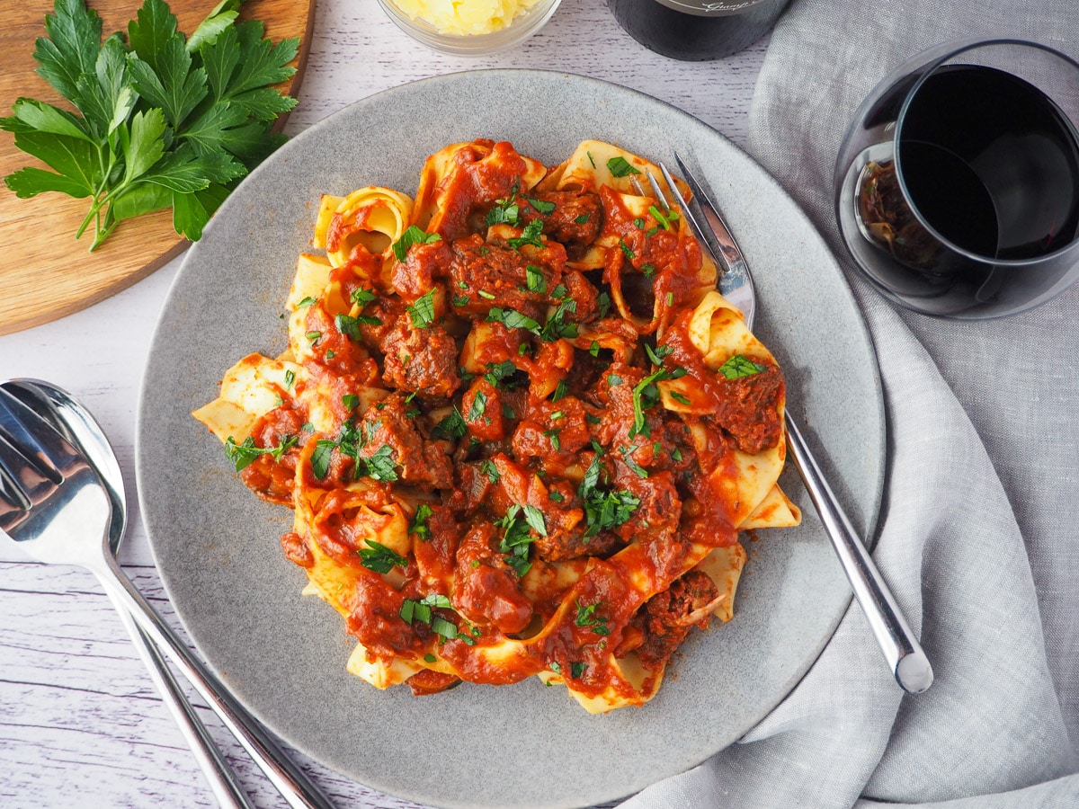 Slow cooker lamb ragu 2 Plate of slow cooker lamb ragu, garnished with chopped parsley, surrounded by serving cutlery, glass of red wine, bowl of parmesan cheese and chopping board with parsley on it.