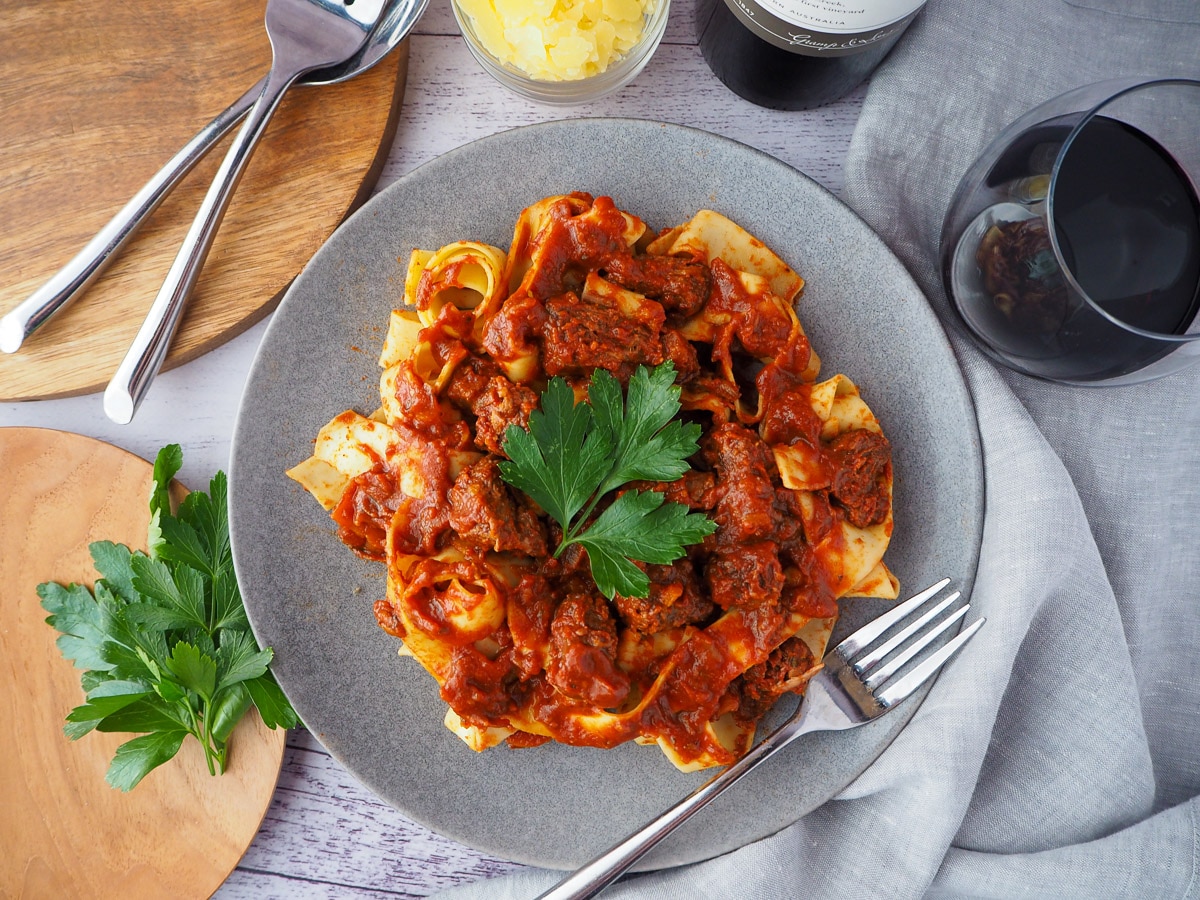 Slow cooker lamb ragu 1 Plate of slow cooker lamb ragu, garnished with parsley, surrounded by serving cutlery, glass of red wine, bowl of parmesan cheese and chopping board with parsley on it.