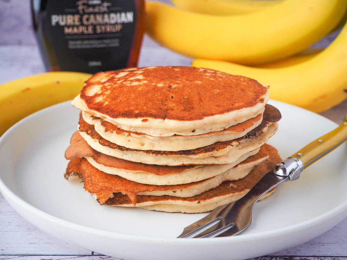 Banana pikelets 15 Stack of banana pikelets on a plate, with a vintage spoon on the side, surrounded by fresh whole bananas and bottle of maple syrup in the background.