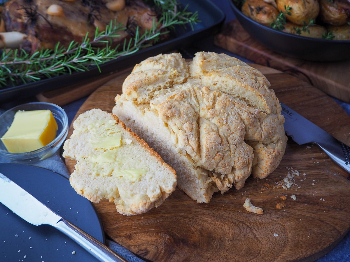 Damper 14 Cut buttered damper on a chopping board, with butter in dish, bread knife and serving dishes with slow cooker lamb shoulder and roasted mini potatoes.