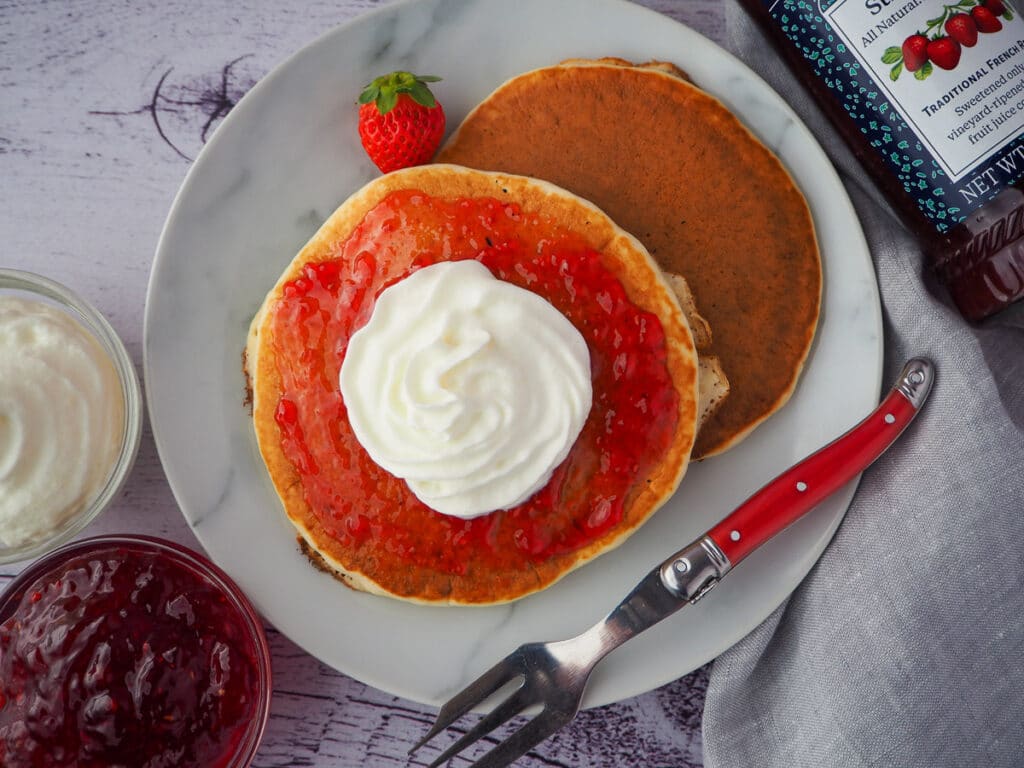 Pikelets 14 Top down view of two pikelets with jam and cream on a plate, with a fresh strawberry, fork, bowl of whipped cream jam and jar of strawberry jam on the sides.