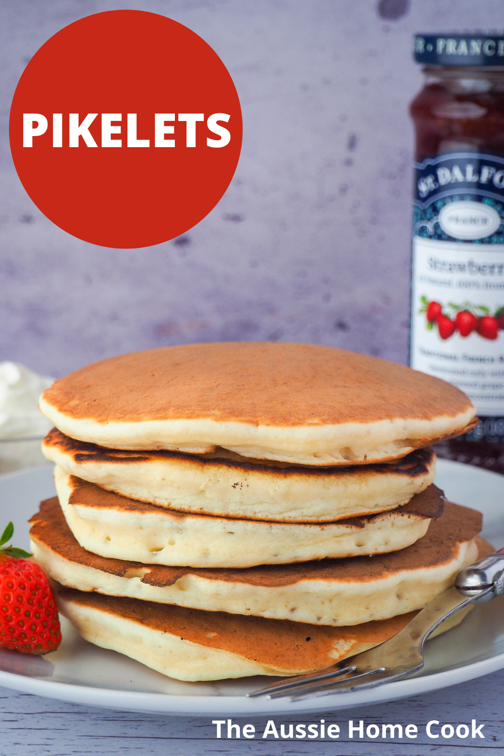 Pikelets 17 Stack of pikelets on a plate, with a fresh strawberry and fork, and whipped cream and jar of strawberry jam in the background, with text overlay Pikelets, The Aussie Home Cook.