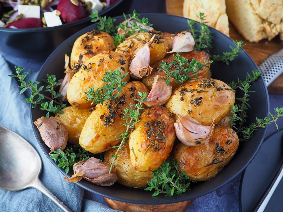 Roasted mini potatoes 2 Roasted mini potatoes in a serving dish garnished with fresh herbs, silver serving spoon to the side, fresh damper, pat of butter, beetroot salad and slow cooker roast lamb on the side.