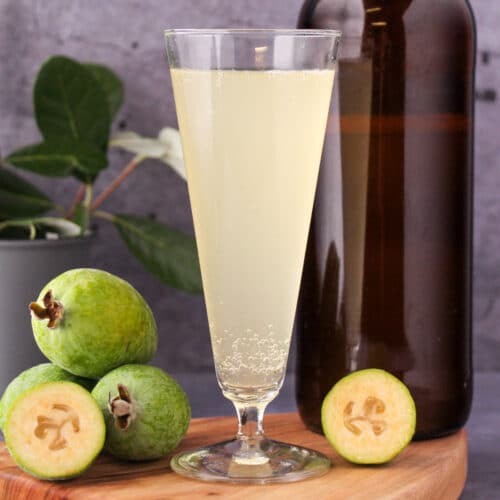 Feijoa fizz 7 Close up tall glass of feijoa fizz on a board, with bottle of feijoa fizz, fresh feijoas and feijoa leaves.