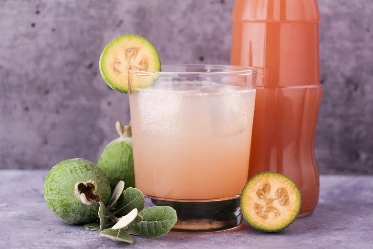 Feijoa syrup 1 Glass of made up feijoa syrup with ice and fresh feijoa garnish, with bottle of feijoa syrup and fresh feijoas and leaves.