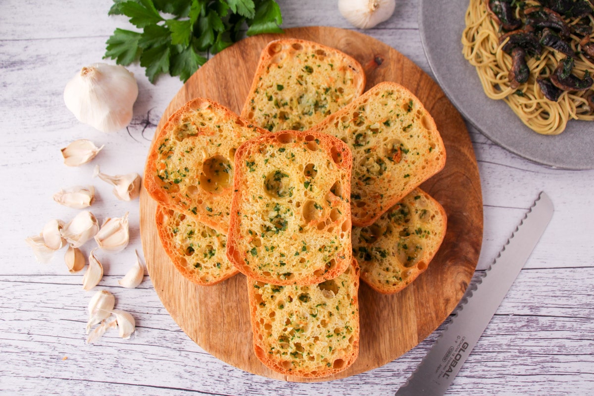 Air Fryer Garlic Bread 11 Stack of air fryer garlic bread on a chopping board, with bread knife, fresh garlic cloves, fresh parsley and a plate of pasta on the sides.