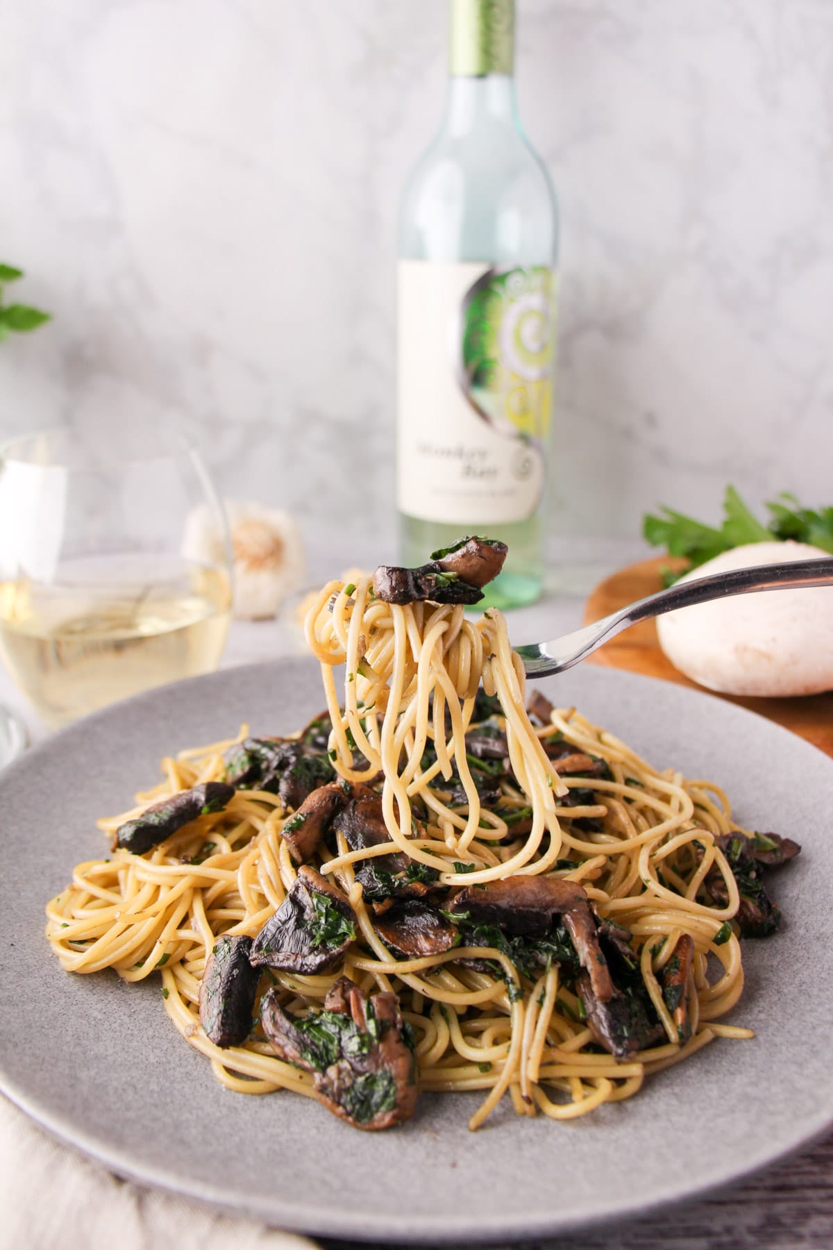 Mushroom Aglio Olio 19 Fork of mushroom aglio olio held over a plate of pasta, with fresh mushrooms, parsley, garlic and a glass and bottle of white wine the background.