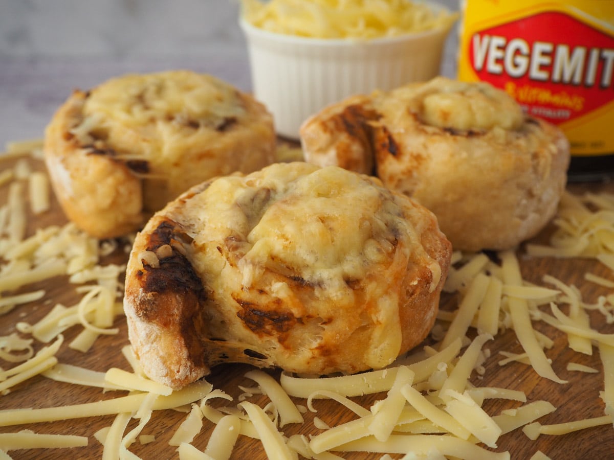 Cheesymite Scrolls 14 Scrolls on a board surrounded by grated cheese, with bowl of cheese and jar of vegemite in the background.