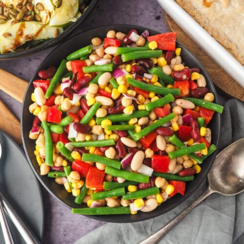 Four Bean Salad 3 Serving bowl of salad with serving spoon, knife, fork and plate on the and pie and rocket and pear salad in the background.