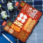 Picnic Charcuterie Board 11 Multi compartment container filled with various charcuterie food, on a picnic blanket with a plate and drinks.