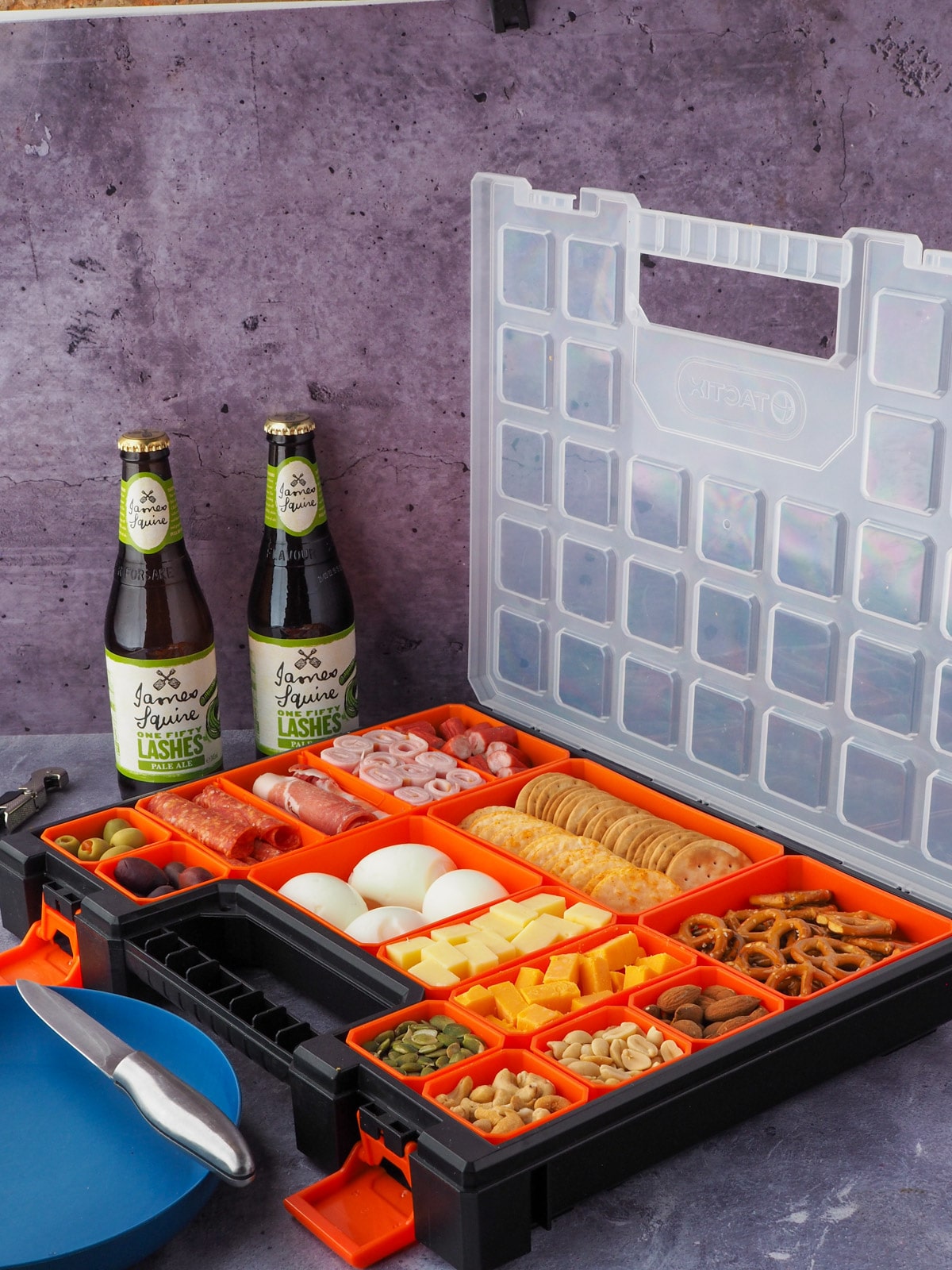 Picnic Charcuterie Board 13 Multi compartment container filled with various charcuterie food, with picnic plate and drinks in the background.