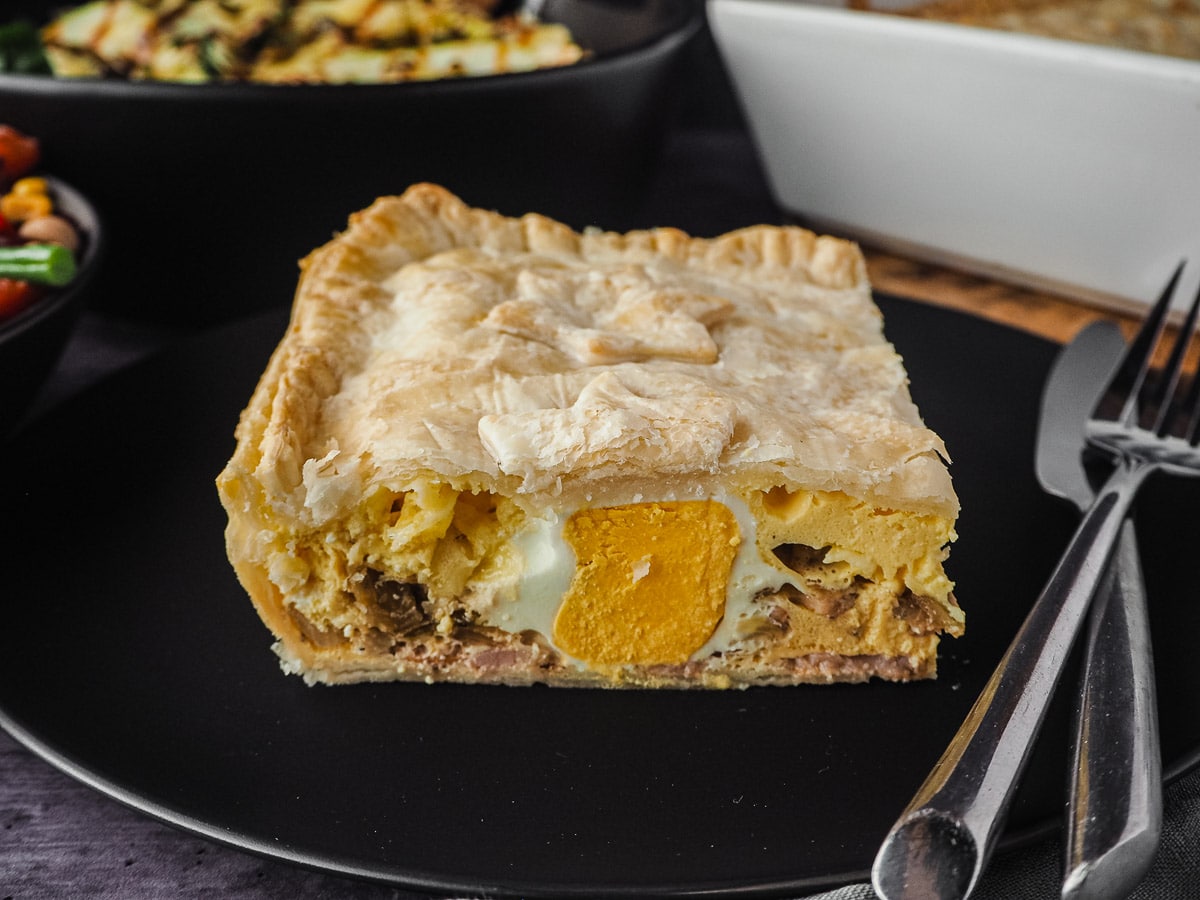 Bacon and Egg Pie 8 Pie in a plate with cutlery, with pie dish and salads in background.