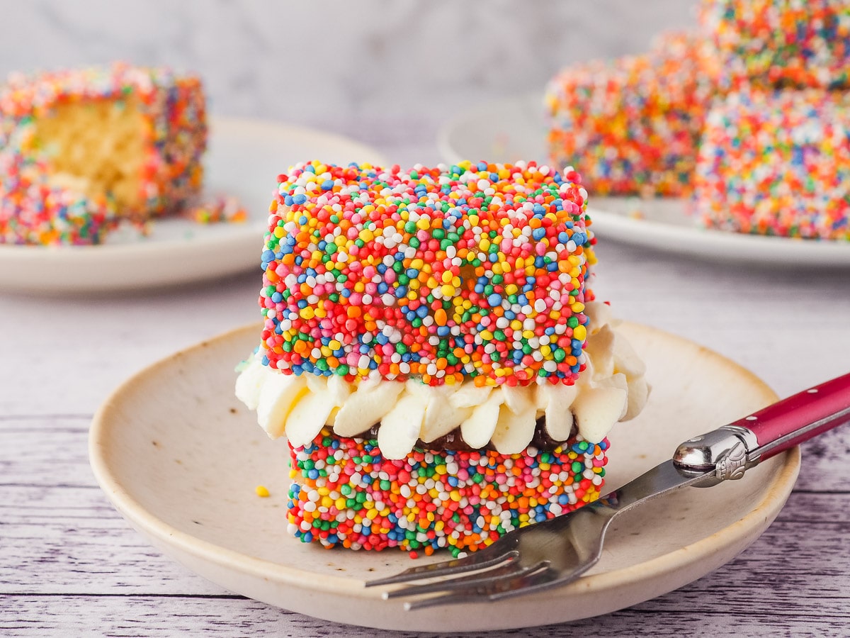Fairy Bread Lamingtons 9 Lamington filled with jam and cream on a plate with a fork and more lamingtons in the background.
