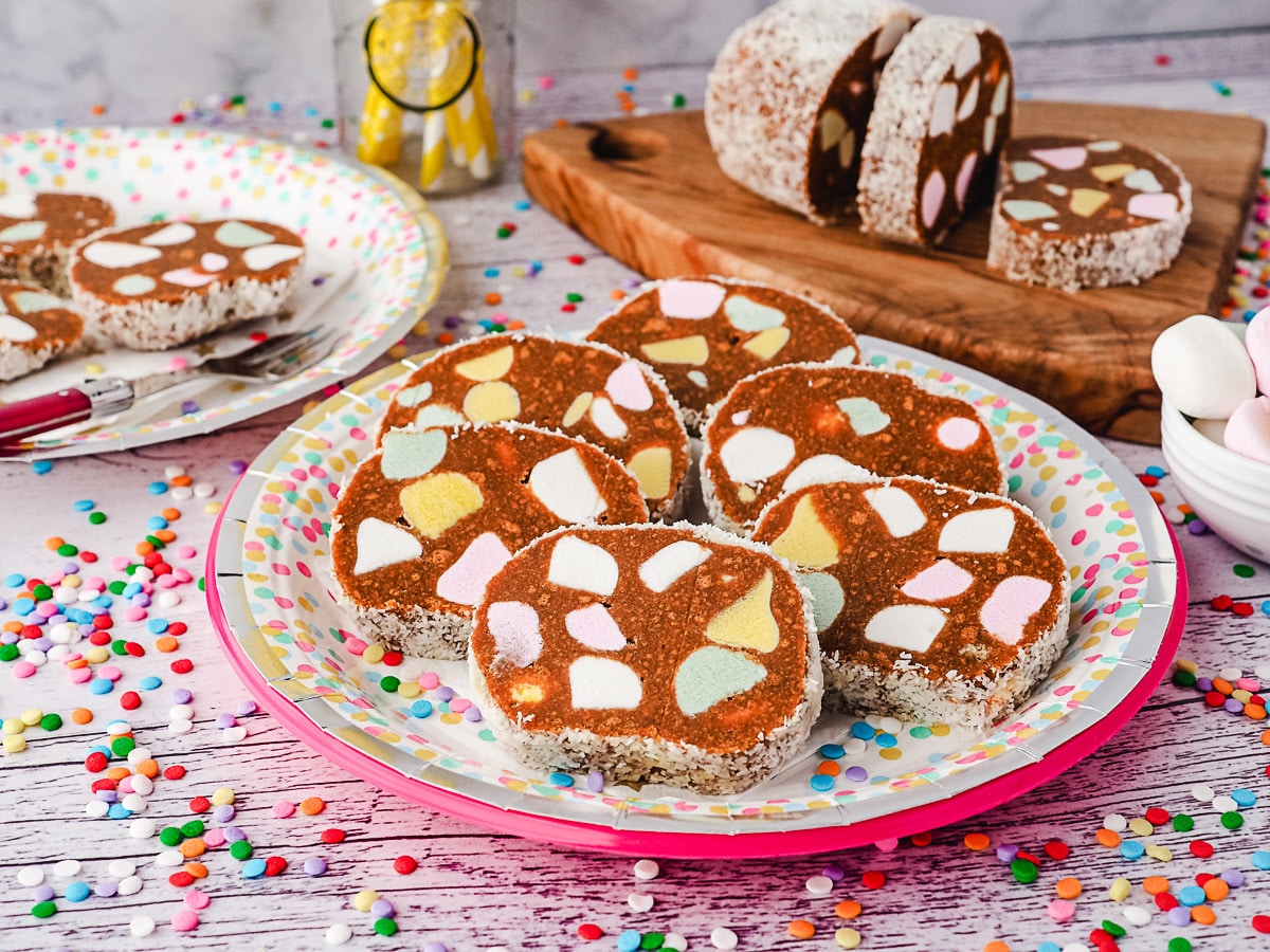 Lolly Cake 7 Slice of lolly cake on a party place, with another slice on a plate, lolly cake log and fruit puffs in background.