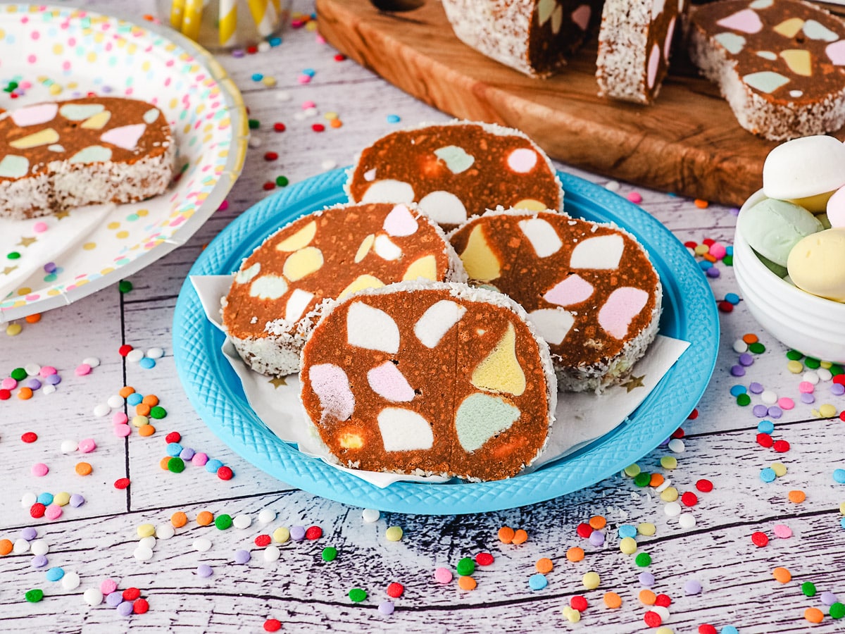 Lolly Cake 1 Slice of lolly cake on a party place, with another slice on a plate, lolly cake log and fruit puffs in background.