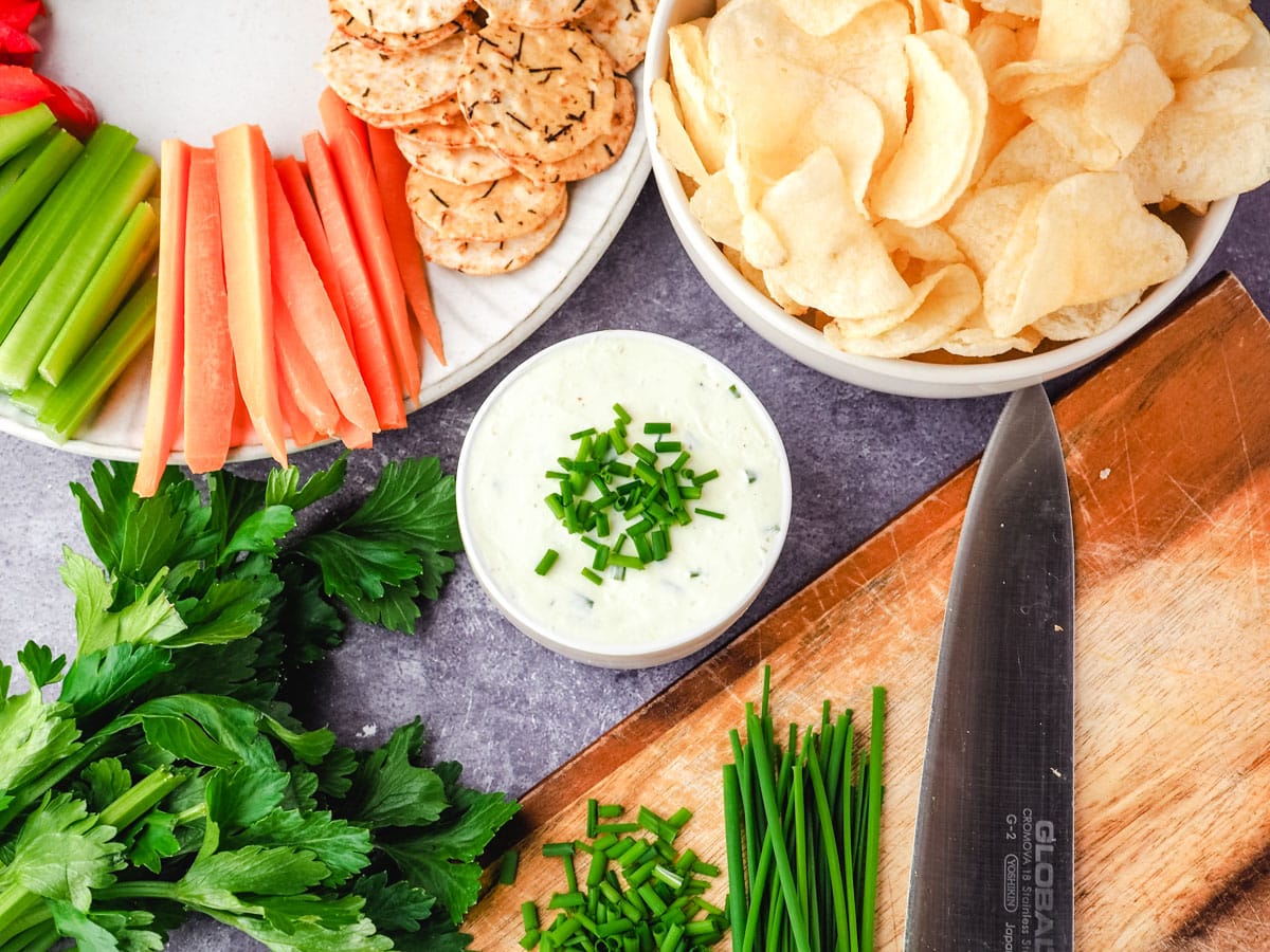 Sour Cream Dip 1 Sour cream dip garnished with fresh chives, with a bowl of chips, plate of crackers and veggie sticks, and chopping board with freshly chopped chives.