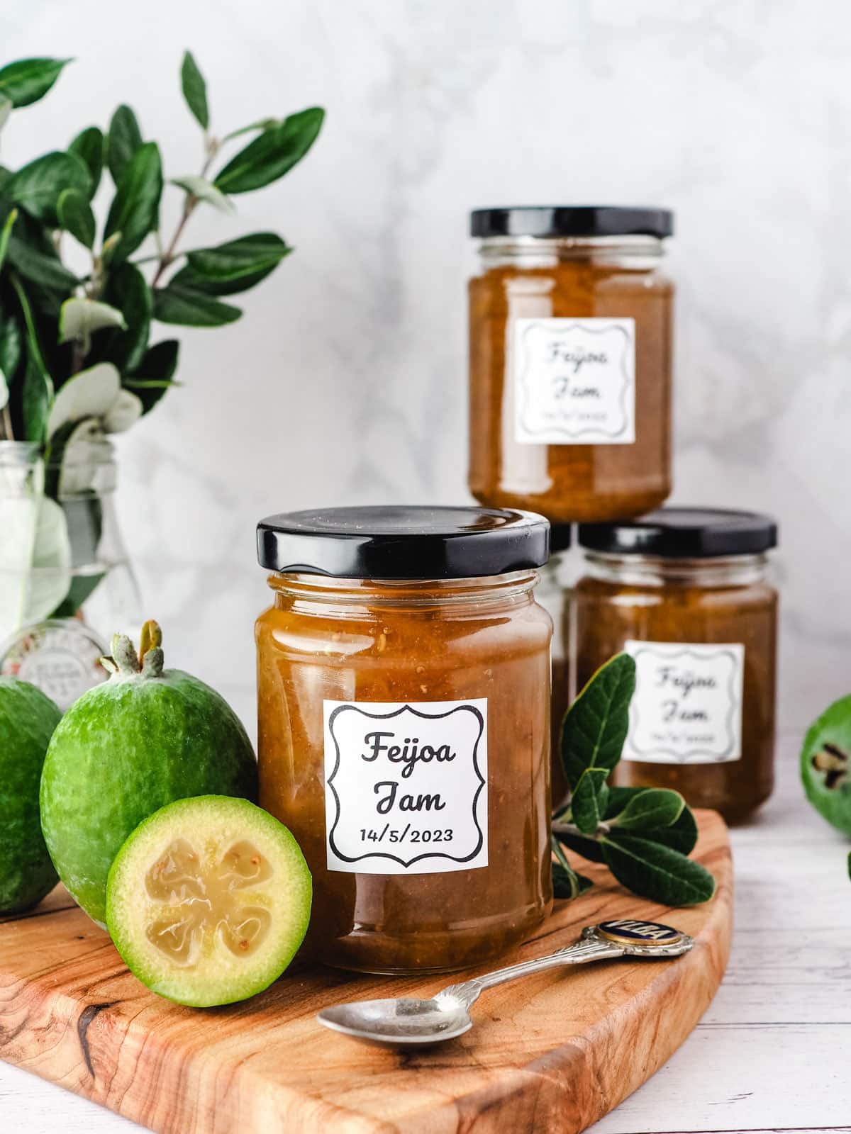 Feijoa Jam 9 Jar of feijoa jam on a serving board with fresh feijoas and leaves on the side, with a stack of feijoa jam jars in the background.