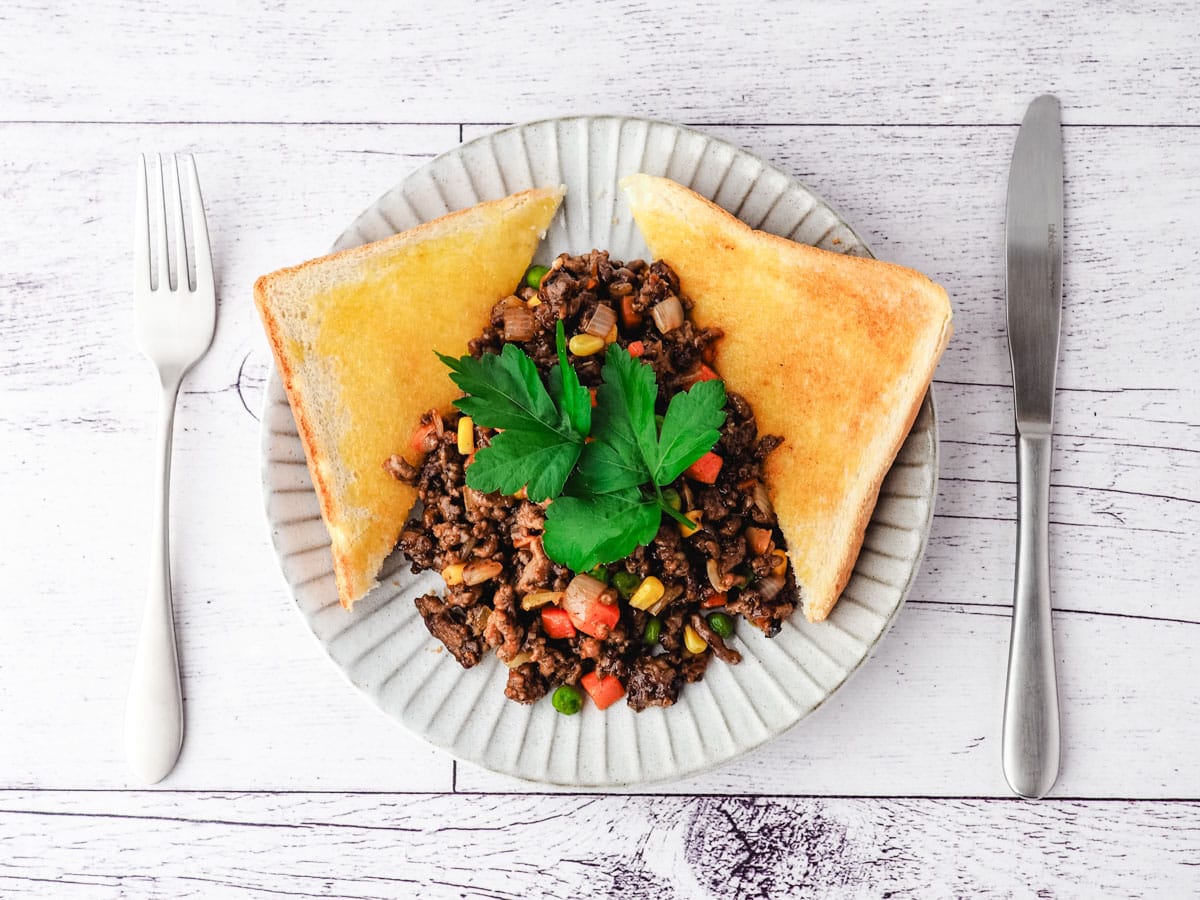 Savory Mince 7 Savory mince on a plate with buttered toast on the side, garnished with flat parsley, with a knife and fork on the side.