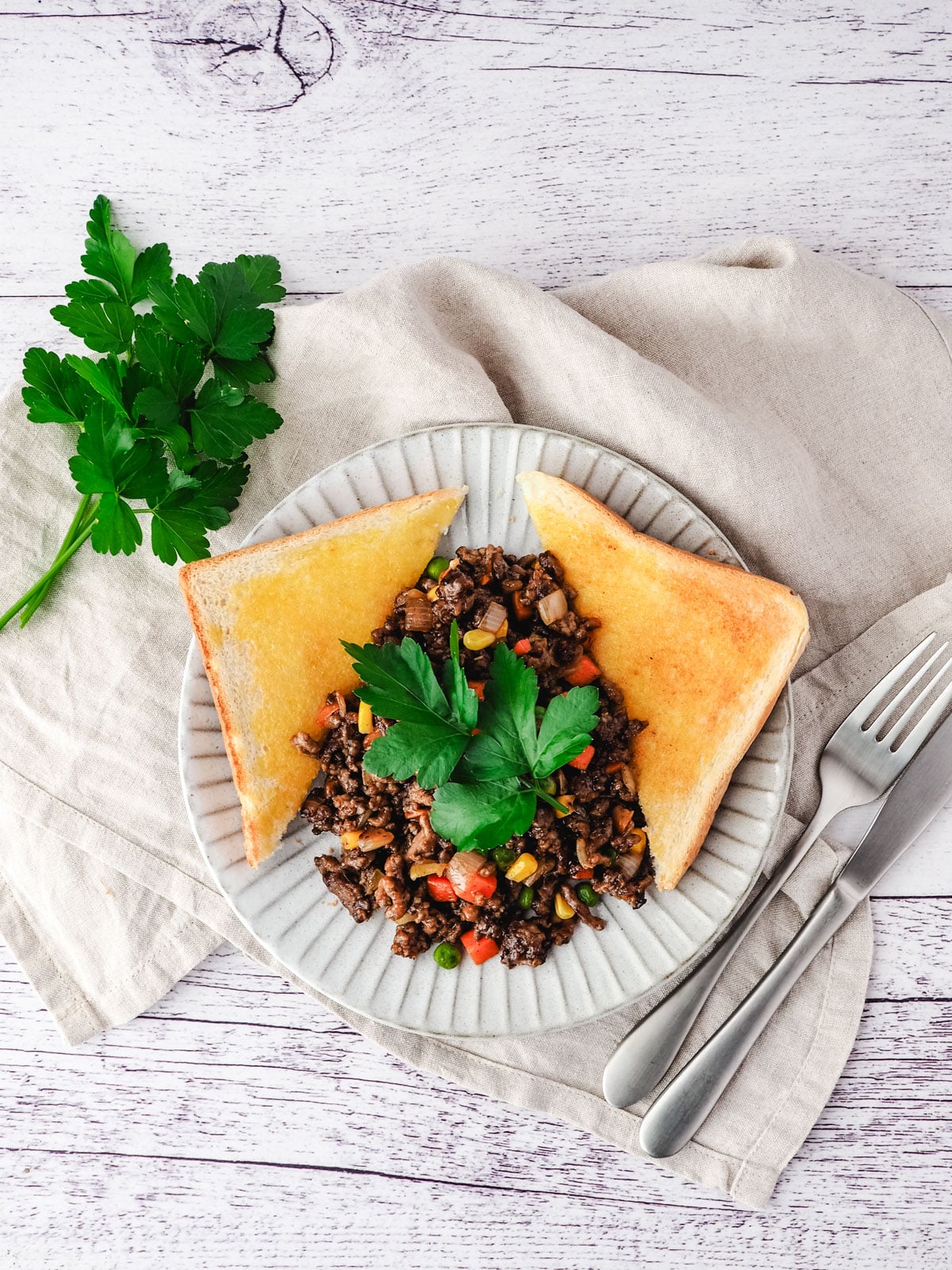 Savory Mince 9 Savory mince on a plate with buttered toast on the side, garnished with flat parsley, with a knife and fork on the side.