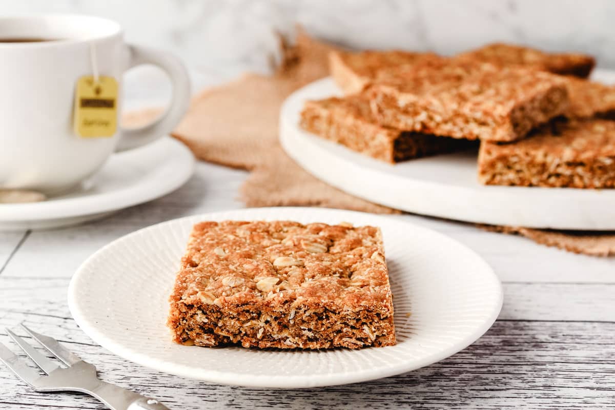 Anzac Slice 7 A piece of Anzac biscuit slice on a plate, with a cup of tea and serving board with rest of pieces of slice in the background.
